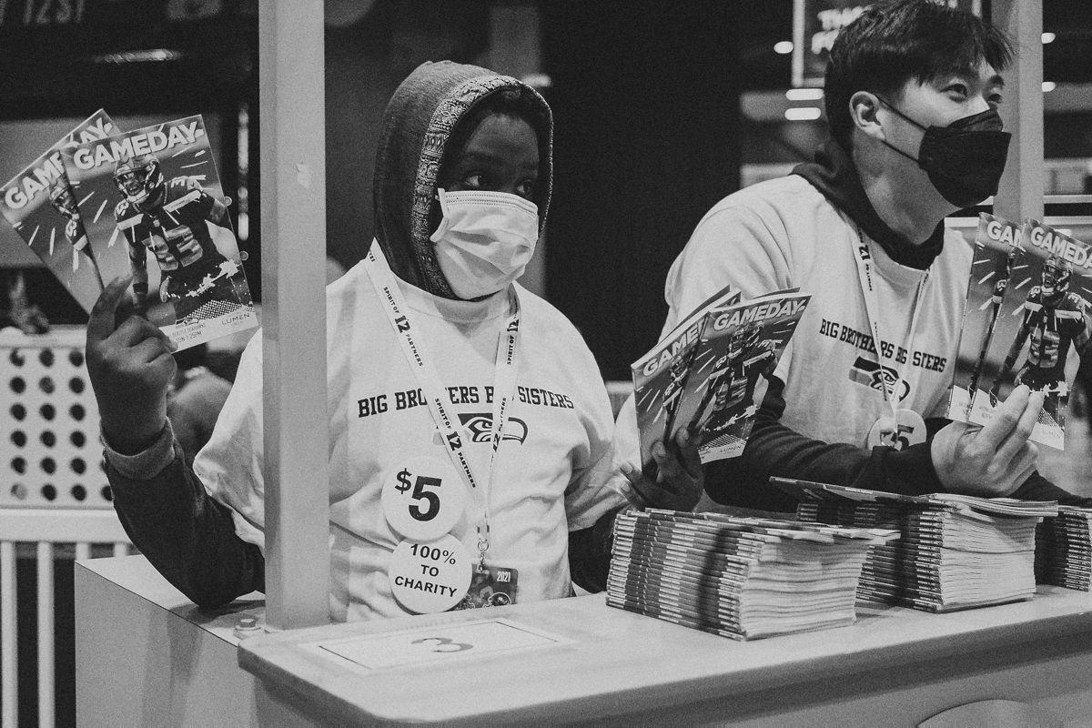 Caleb stands left, masked and holding up programs to sell. Austin stands t his right, also masked, leaning over the booth talking to someone out of frame.